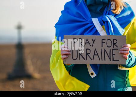 Prega per l'Ucraina. Donna coperta da bandiera ucraina che tiene anti guerra segno cartone all'aperto. Monumento a croce religiosa defocused Foto Stock