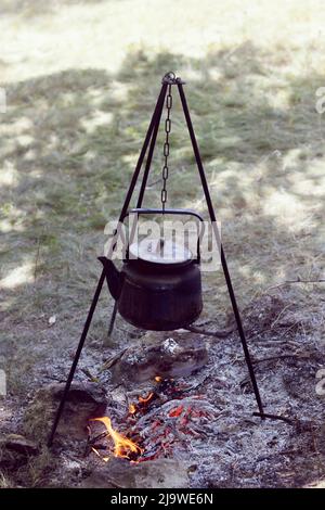 I viaggiatori scaldano l'acqua in un bollitore nero su un fuoco dai rami della foresta. La prima colazione del turista in natura Foto Stock