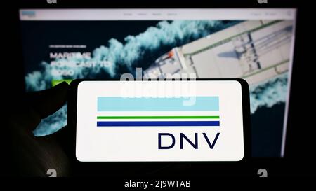 Persona che detiene il telefono cellulare con il logo della società di servizi norvegese DNV SULLO schermo di fronte alla pagina web aziendale. Mettere a fuoco sul display del telefono. Foto Stock