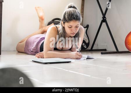 donna latina con capelli bianchi dipinte, giacente sul pavimento, profondamente in pensiero, scrivendo su un quaderno gli obiettivi e gli obiettivi della sua dieta quotidiana Foto Stock