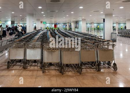 Barcellona, Spagna - 15 aprile 2022 - Vista di molti carrelli bagagli aeroporto vuoti accatastati in fila nella sala del terminal dell'aeroporto con i passeggeri in attesa Foto Stock