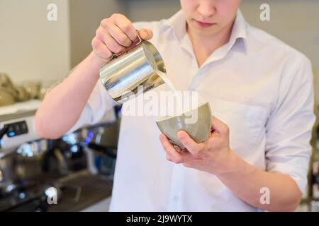Primo piano di un giovane barista che versa il latte in una tazza per preparare un caffè fresco in un bar Foto Stock