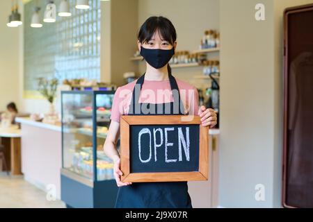 Cameriera asiatica in maschera in piedi al bar con un cartello aperto durante la pandemia di coronavirus Foto Stock