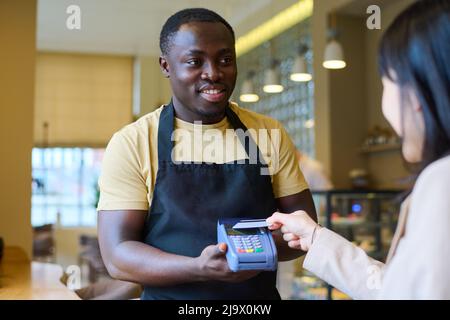 Giovane cameriere africano in uniforme tenendo terminale e sorridendo mentre il cliente paga con carta di credito dopo pranzo al ristorante Foto Stock