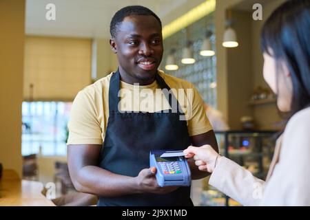 Giovane cameriere africano in uniforme tenendo terminale e sorridendo mentre il cliente paga con carta di credito dopo pranzo al ristorante Foto Stock