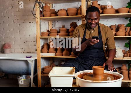 vasaio misto da corsa afro maschio con grembiule nero ed elegante camicia scura seduta al tavolo da officina vasaio con vaso in argilla marrone fatto a mano Foto Stock