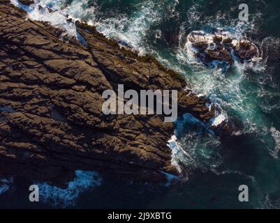 Una vista aerea di una grande pietra ruvida contro la quale l'acqua blu dell'oceano batte con onde bianche e schiumose. Bella stagcape, scene calme. Bellezza Foto Stock