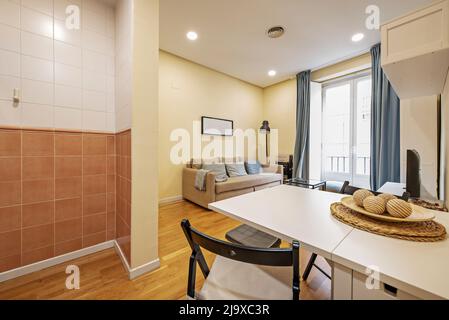 Appartamento monolocale con tavolo da pranzo bianco pieghevole con sedie nere abbinate, divano letto a tre posti rivestito in tessuto beige e balcone con vetro Foto Stock