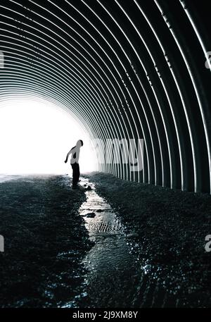 Bambino in piedi vicino a ruscello che corre attraverso tunnel di metallo scuro. Foto Stock