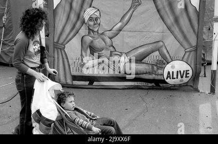 Una donna con un bambino in un passeggino passa accanto a un poster circense raffigurante un uomo sdraiato su un letto di unghie. Foto Stock