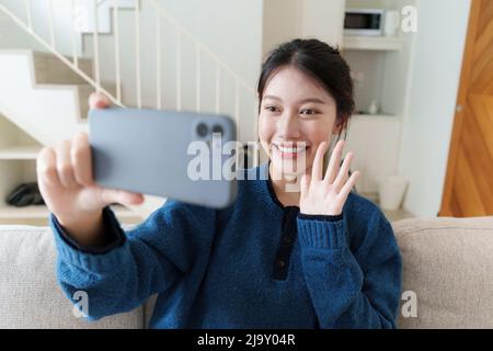 Ritratto di giovane donna asiatica videochiamata con la sua amica e controllare i social media da smartphone seduto sul divano. Concetto di stile di vita. Foto Stock