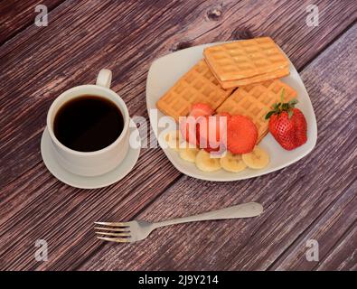 Waffle freschi viennesi con fragole e banane su un piatto, accanto a una tazza di caffè nero su un tavolo di legno. Vista dall'alto Flat Lay. Foto Stock