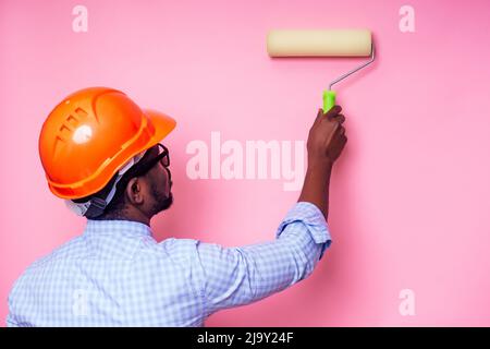 Nero uomo Africano americano tenendo rullo di vernice in mano dipinge la parete in colore rosa .Happy africano costruttore pittura all'interno della casa, uomo d'affari Foto Stock