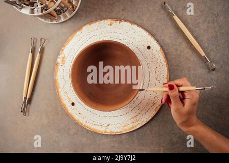 giovane e attraente vasaio femminile che lavora in uno studio con un vaso di argilla marrone nel laboratorio di artigianato tenendo l'attrezzo e la tazza in mano . business Foto Stock