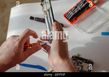 Repair of smartphones concept. A man unscrews the screws fixing the display from the board. Part of a series. Inside the room. Selective focus. Foto Stock