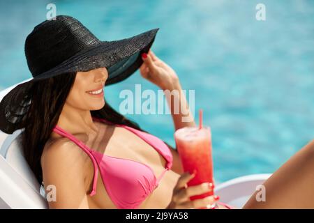 Bella brunetta che riposa sul lettino, nascondendo gli occhi sotto la sposa di cappello nero all'aperto. Vista laterale di affascinante femmina in bikini rosa che beve frullato fresco, mentre si sdraia sul lettino solare al resort. Concetto di riposo. Foto Stock