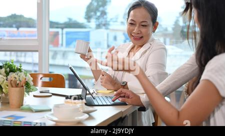 Leader positivo del team femminile di mezza età che spiega i dettagli del progetto ai giovani dipendenti durante la riunione Foto Stock