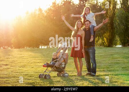 Felice fotosessione attiva di famiglia nel parco estivo. Bella madre dai capelli lunghi suo marito con sua figlia e figlio in un passeggino riposano sul Foto Stock