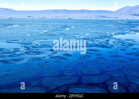 Un'ora blu d'inverno vista mattutina attraverso la laguna di Jökulsarlön iceberg senza ghiaccioli presenti. La laguna è coperta da uno strato rotto di ghiaccio sottile. Foto Stock