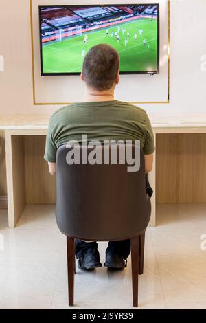 Un uomo solista si siede scomodo in una sedia e guarda una partita di calcio sulla televisione appesa al muro della stanza. Foto Stock