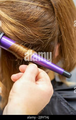 L'acconciatore ricci acconciature di capelli lunghi marrone con il ferro arricciacapelli in parrucchiere, primo piano Foto Stock