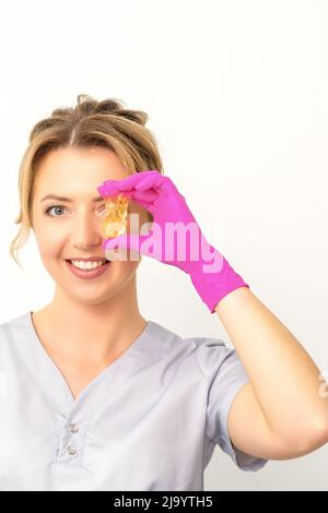 Il padrone di rimozione dei capelli dello zucchero tiene la pasta gialla liquida dello zucchero, coprente il suo occhio con cera per depilazione su uno sfondo bianco Foto Stock