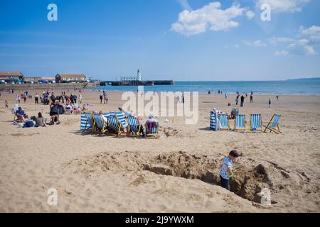 Un ragazzino in primo piano scavando un grande buco nella sabbia mentre i gruppi di famiglia si siedono in sdraio dietro a Scarborough, North Yorkshire Foto Stock