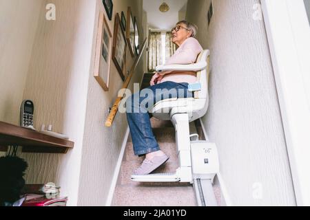 Donna anziana che usa il sollevamento automatico della scala su una scala a casa sua. Ascensore medico per disabili e anziani in casa. Selettivo per Foto Stock