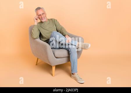 Foto a tutta lunghezza di funky anziano grigio hairdo uomo sit indossare occhiali pullover scarpe jeans isolato su sfondo beige Foto Stock