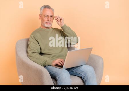 Foto di rigido anziano grigio hairdo uomo sit lavoro laptop indossare occhiali pullover jeans isolato su sfondo beige Foto Stock