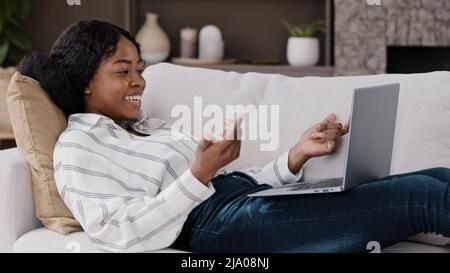 Donna africana sdraiata sul divano a casa guardare lo schermo del laptop saluto hello fare videochiamata parlando avendo conversazione remota con amici che vivono in famiglia Foto Stock
