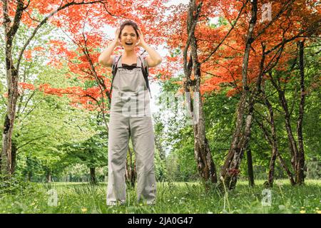 la ragazza adolescente urla, esprime emozioni, è sollecitata dal rumore, le mani che coprono le orecchie, contro sfondo di bella natura, da sola nella foresta Foto Stock