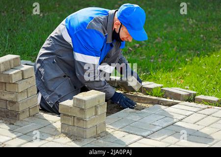 Il muratore in abiti da lavoro si siede sul marciapiede e stende le lastre di pavimentazione. Vista dell'uomo in attività all'aria aperta. Costruttore professionista fa organizzazione del territorio giorno d'estate. Scena reale. Foto Stock