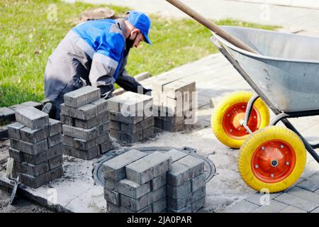 Il muratore in abiti da lavoro si siede sul marciapiede e stende le lastre di pavimentazione. Vista dell'uomo in attività all'aria aperta. Costruttore professionista fa organizzazione del territorio giorno d'estate. Scena reale. Foto Stock