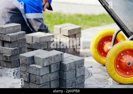 Il muratore in abiti da lavoro si siede sul marciapiede e stende le lastre di pavimentazione. Vista dell'uomo in attività all'aria aperta. Costruttore professionista fa organizzazione del territorio giorno d'estate. Scena reale. Foto Stock