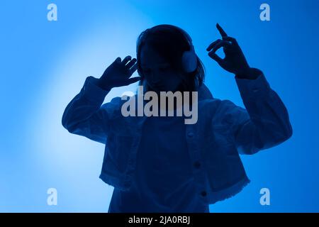 Teen girl in eleganti cuffie danze alla musica in cuffie bianche su sfondo studio in trendy 80s 90s club blu festa luce. Foto Stock