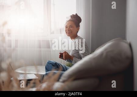 Bella giovane donna di razza mista di buon umore che si rilassa sul divano in casa accogliente con una tazza di tè Foto Stock