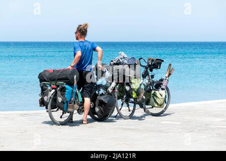 Un uomo si riposa e si alza reggendo due biciclette mentre guarda verso il mare. Le biciclette sono molto cariche di equipaggiamento per una vacanza o un tour in bikepacking Foto Stock