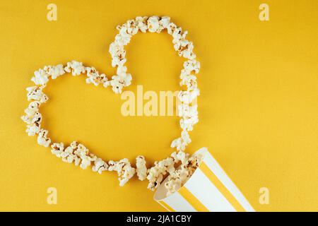 popcorn sparso da una tazza a strisce di carta su sfondo giallo a forma di cuore. il concetto di home cinema e film nel cinema Foto Stock