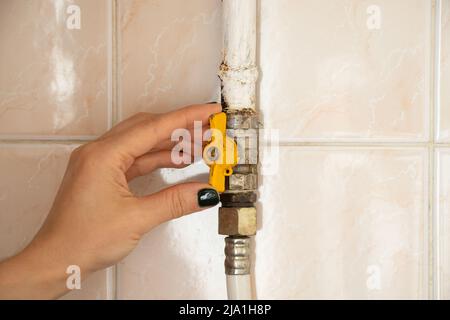 La mano chiude il rubinetto sul tubo del gas contro lo sfondo delle piastrelle della casa in appartamento, il rubinetto sul tubo del gas nel cucinotto Foto Stock