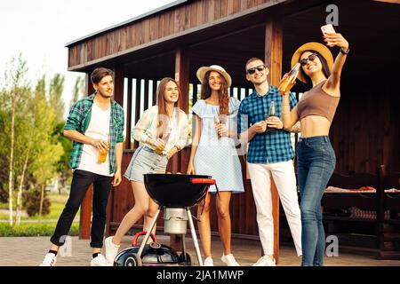 Un gruppo di giovani amici allegri che hanno una festa barbecue nel cortile, prendere un selfie su un telefono cellulare, grigliare carne, fare toast, bere le Foto Stock