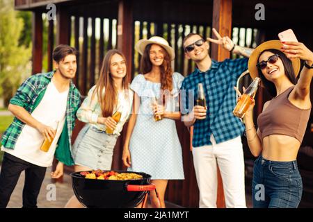 Un gruppo di giovani amici allegri che hanno una festa barbecue nel cortile, prendere un selfie su un telefono cellulare, grigliare carne, fare toast, bere le Foto Stock