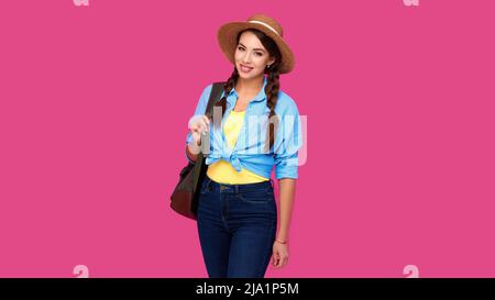 Viaggiatore positivo donna in abiti casual, cappello di paglia e zaino su sfondo rosa isolato. Giovane turista caucasico sorridente Foto Stock
