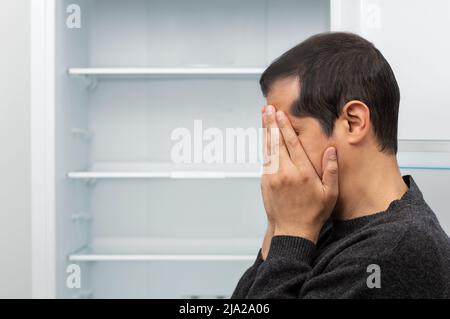 Primo piano di un uomo che copre il suo volto e piangendo con un frigorifero vuoto sullo sfondo in concetto di crisi economica e spazio copia Foto Stock