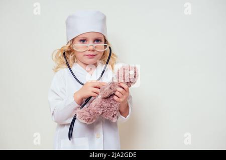 Adorabile bambina in uniforme indossando il cappuccio del medico e gli occhiali che giocano il medico e l'orsacchiotto di ascolto con stetoscopio su sfondo bianco.capretto femmina Foto Stock