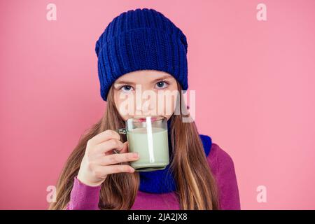 Bella ragazza con un bicchiere di latte in un cappello a maglia in studio su uno sfondo rosa.ragazza bambino felice bere yogurt notte di natale Foto Stock
