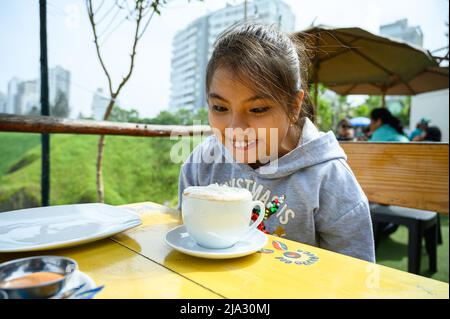 Bella ragazza seduta nel bar con una tazza di cioccolata calda con panna montata che lo guarda felicemente Foto Stock