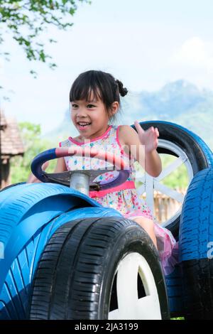 La ragazza piccola carina che si diverte nel parco giochi all'aperto. Una bambina corre su un'auto giocattolo fatta di vecchi pneumatici nel parco. Sana attività estiva per chil Foto Stock