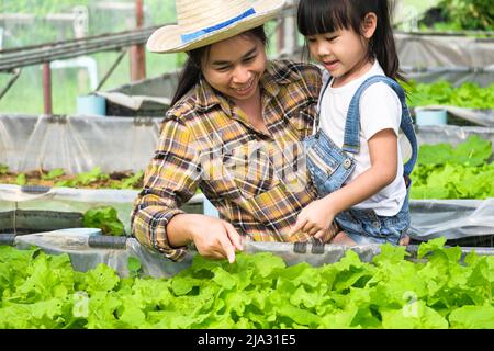 Madre giovane in cappello di paglia che insegna le sue figlie nel giardino del cortile. La bambina aiuta la madre nel giardino, un piccolo giardiniere. Cute ragazza piantando Foto Stock