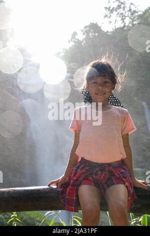 Cute Asian ragazza siede su un ceppo vicino Wachirathan Waterfall, guarda la macchina fotografica e sorrisi nel Parco Nazionale di Inthanon, Chiang mai, Thailandia. Foto Stock